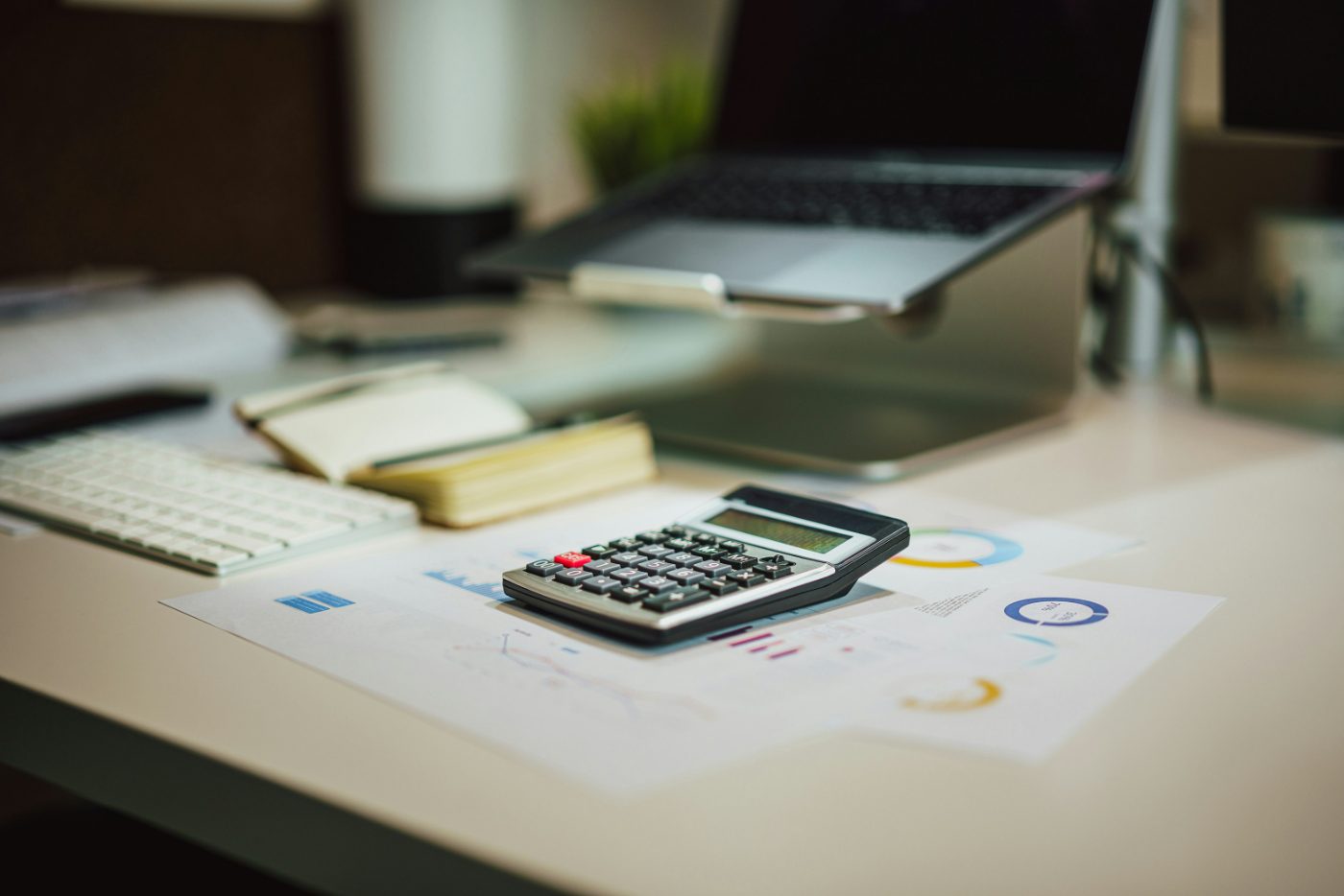 A calculator resting on printed charts on a desk, with a laptop on a stand behind it.