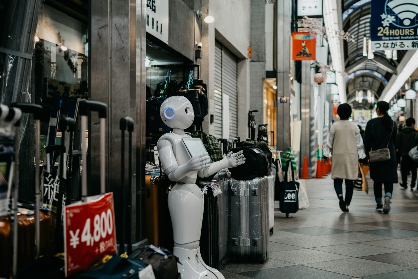 A white humanoid service robot standing beside stacked suitcases in a covered shopping arcade, with shoppers walking past storefronts in the background.