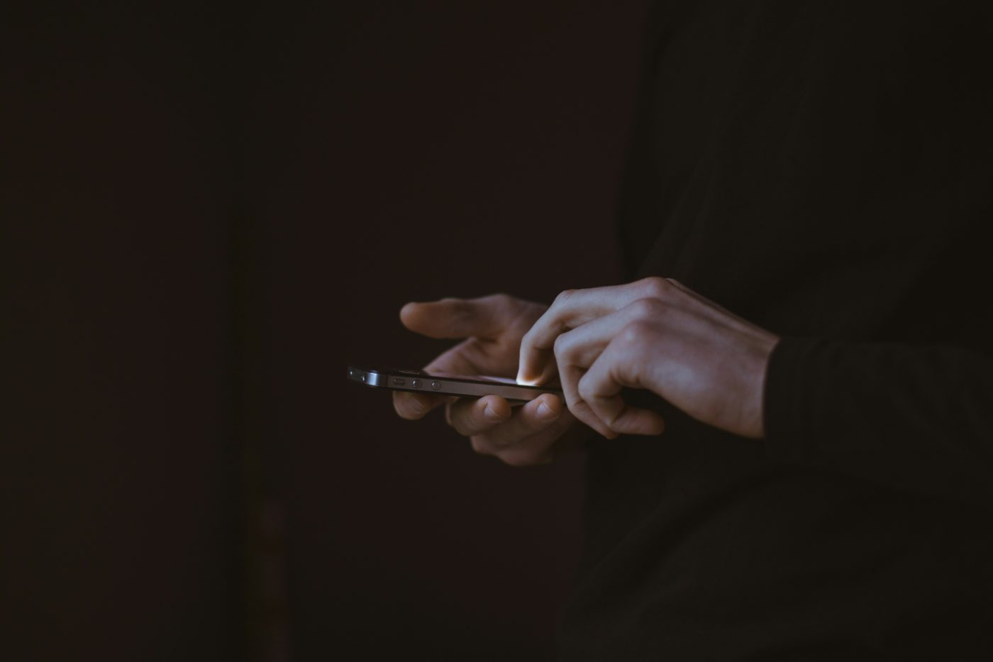 Two hands holding a smartphone in a dark room, one finger tapping the lit screen.